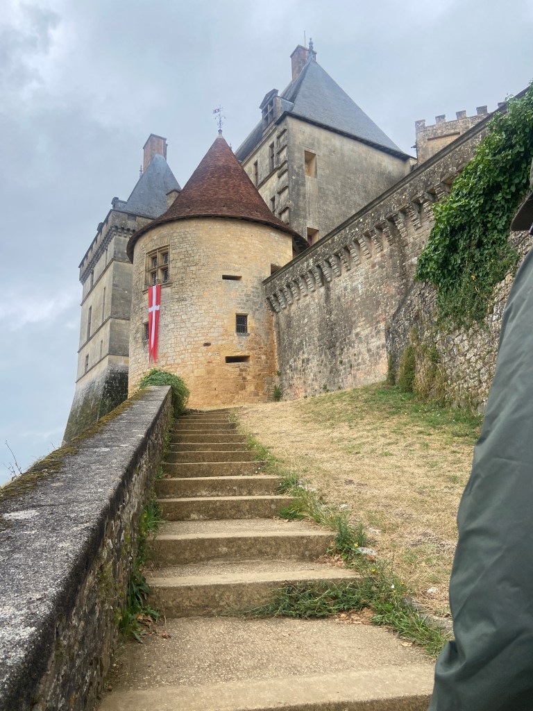 Stairs leading up to a historic stone castle with a red and white flag, surrounded by greenery and overcast skies.