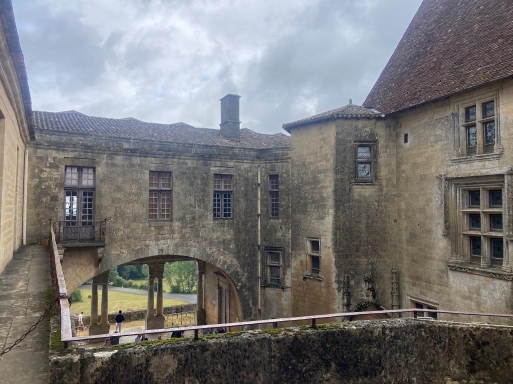 An ancient stone courtyard of a castle, featuring arched windows and a balcony, with a cloudy sky overhead.