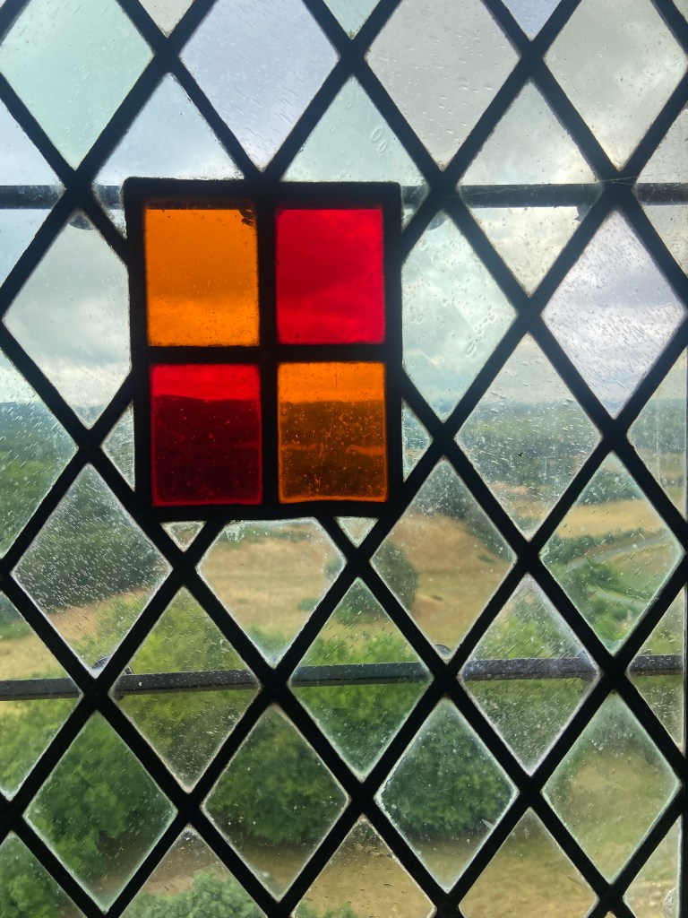 Close-up of a stained glass window featuring a red and orange geometric design, with rainfall droplets visible on the glass and a view of green fields in the background.