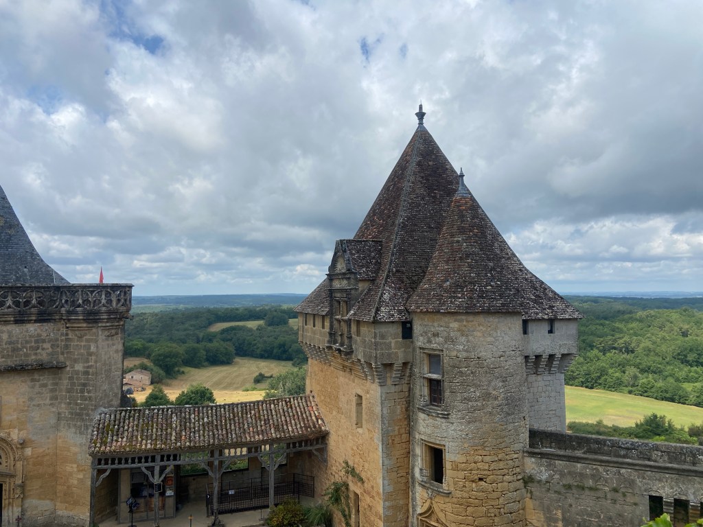 A view of a medieval castle with distinctive towers and a stone structure, surrounded by green fields and trees under a cloudy sky.