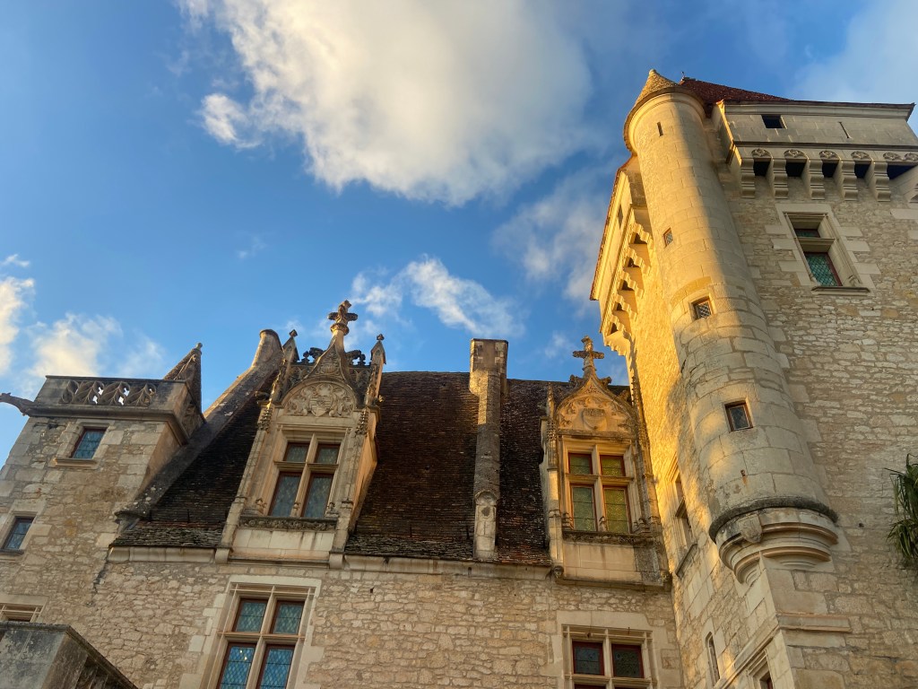 View of a historic castle with ornate architectural details, featuring a tower and steeply pitched roofs against a blue sky with clouds.