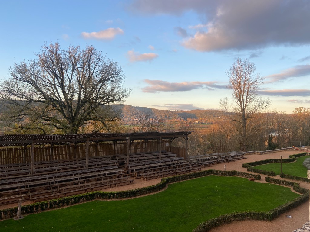 A scenic view of an outdoor amphitheater with wooden benches, surrounded by green grass and trees. The background features rolling hills and a cloudy sky during sunset.