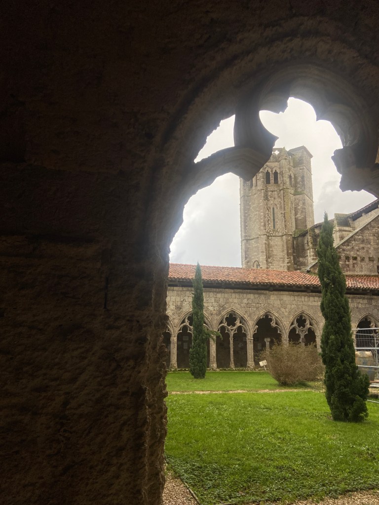 View of a stone cloister through an arched window, featuring a lush green courtyard and a tall medieval tower in the background.