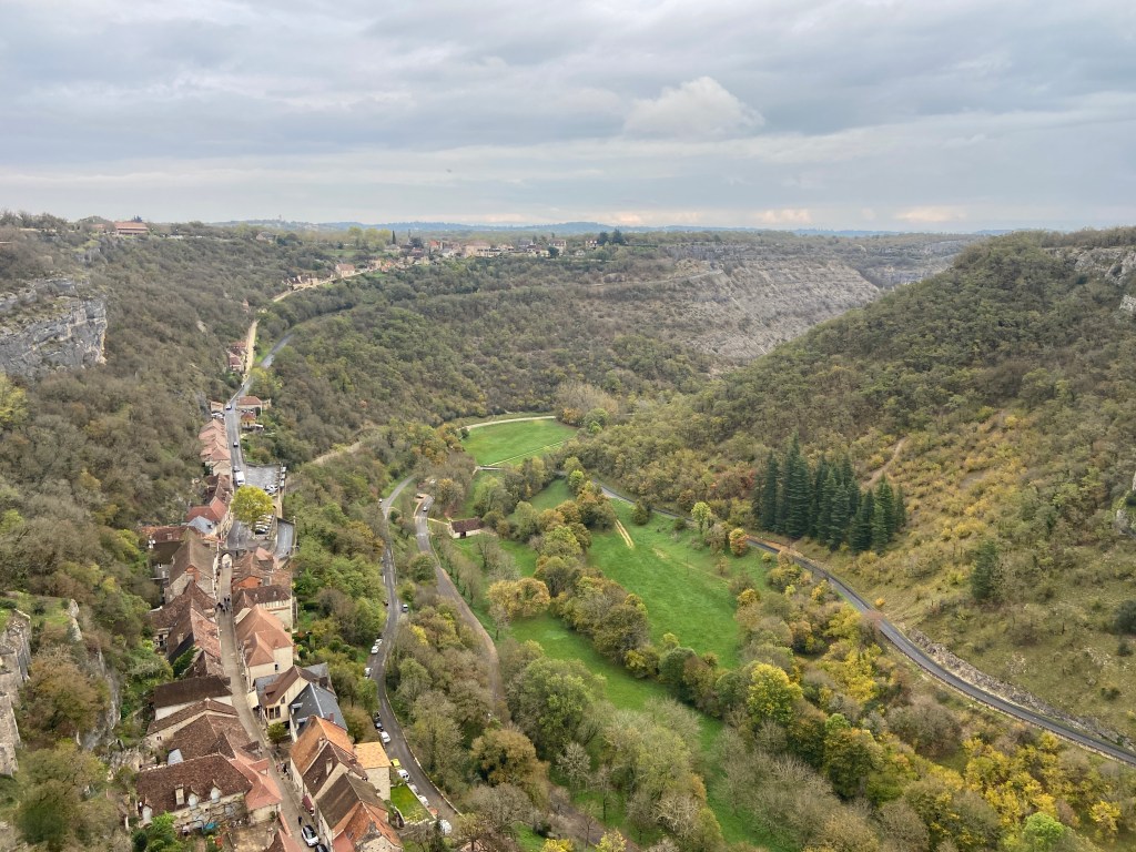 A panoramic view of a lush green valley surrounded by hills, featuring a winding road and a small village with traditional houses situated on the valley's edge.