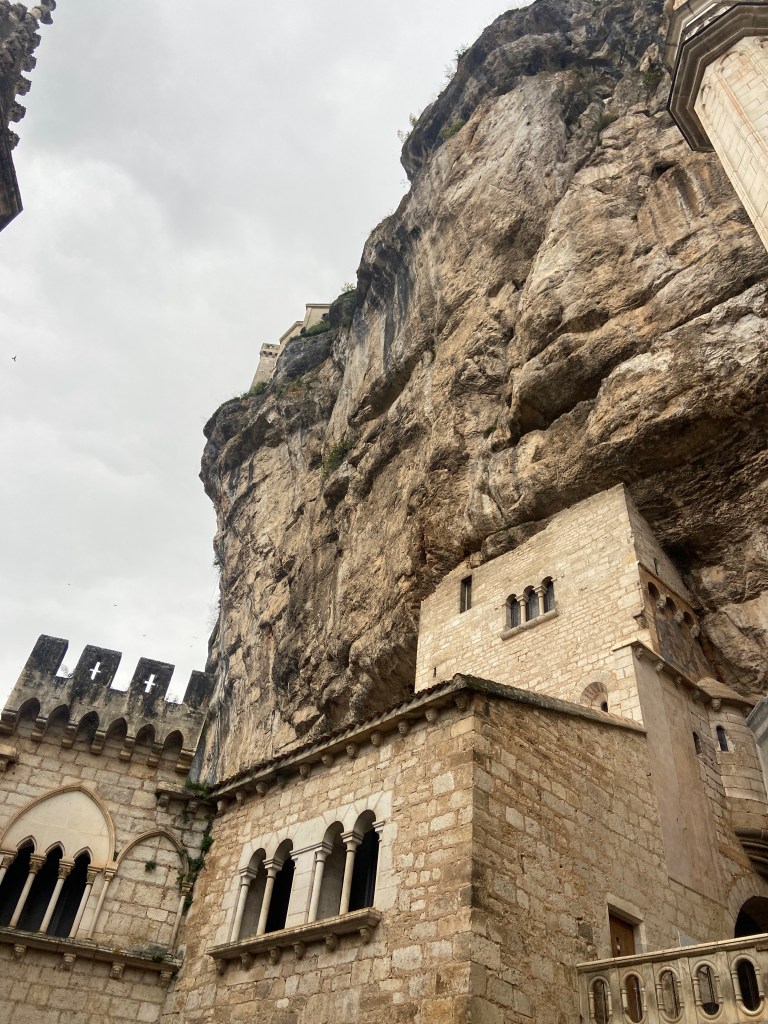 A view of a stone building at the base of a towering rocky cliff under a cloudy sky.
