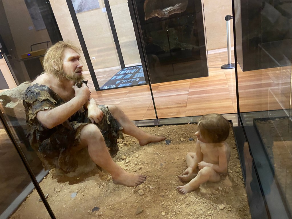 A life-size exhibit depicting a prehistoric man sitting on the ground next to a young child, both surrounded by a sandy environment within a glass display case.