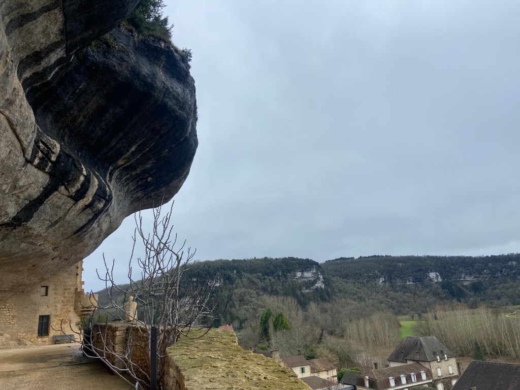 View of a rocky cliff overhanging a landscape with trees and buildings below, under a cloudy sky.