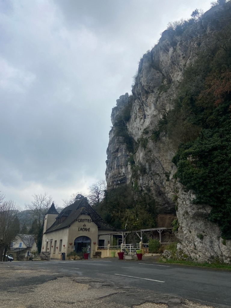 A stone building labeled 'Grottes de Lacave' nestled at the base of a steep cliff under a cloudy sky.